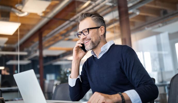 Homem sorridente com o telefone celular junto ao ouvido enquanto olha para um laptop