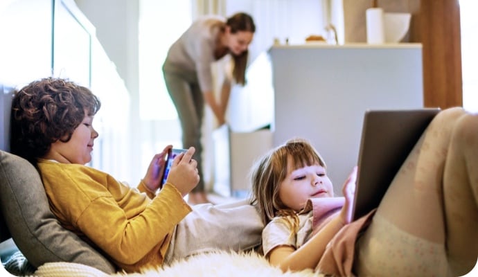 Crianças deitadas com dispositivos tablet enquanto a mãe ao fundo está arrumando a cozinha.