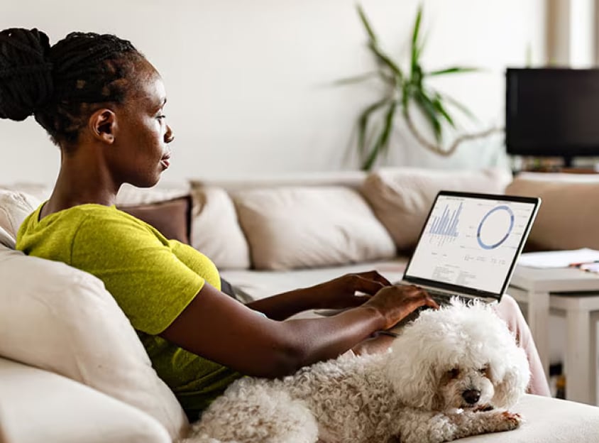Mulher sentada, trabalhando no notebook e com um cachorro ao lado dela.